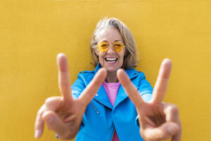 Glückliche reife Frau gestikuliert Friedenszeichen vor auf gelber Wand - OIPF02646