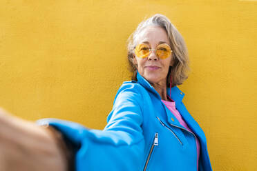 Smiling mature woman taking selfie in front of yellow wall - OIPF02647