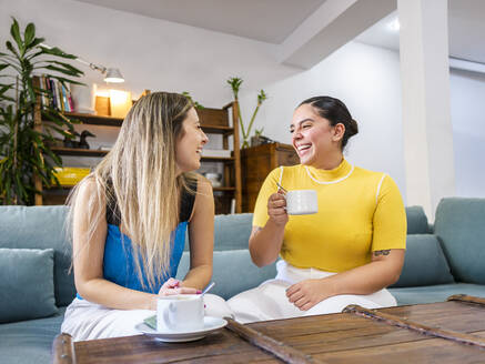 Glückliche Frau mit Kaffeetasse im Gespräch mit einem Freund auf dem Sofa zu Hause - AMRF00106