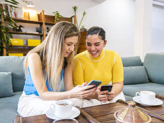 Happy young women sharing mobile phones on sofa at home - AMRF00110