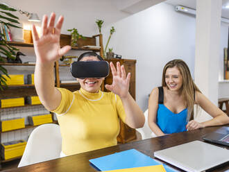 Smiling businesswoman gesturing with VR goggles sitting by friend at table - AMRF00120