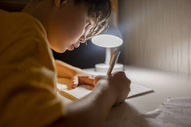 Junge schreibt in Buch unter beleuchteter Lampe zu Hause - ANAF00308