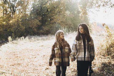 Girl holding hands with sister and walking in forest on sunny day - YLF00024