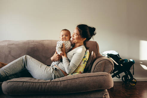 Junge Frau auf einem Sofa liegend mit ihrem Säugling im Arm, lächelnde Mutter, die sich zu Hause um ihr Baby kümmert. - JLPSF21650