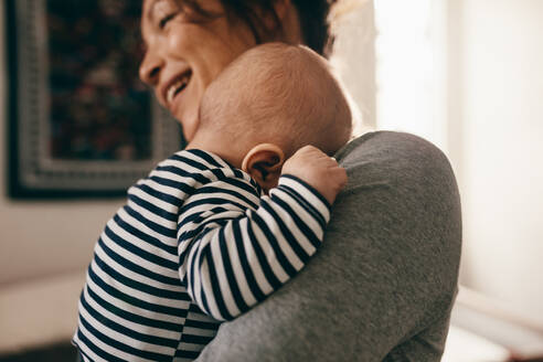 Seitenansicht einer lächelnden Frau, die ihr schlafendes Baby trägt. Das Baby schläft mit seinem Kopf auf der Schulter seiner Mutter zu Hause. - JLPSF21661