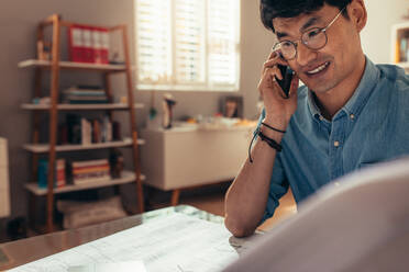 Happy architect looking at drawing and talking over cell phone at his home office. Architect answering phone call at his workplace. - JLPSF21669