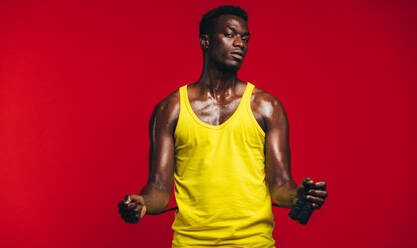 Fit junger Mann mit Springseil auf rotem Hintergrund. Afrikanisches Fitness-Männermodell mit Springseil schaut in die Kamera. - JLPSF21675