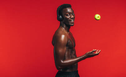 Selbstbewusster junger, muskulöser Mann ohne Hemd und mit Kopfhörern, der einen Apfel vor rotem Hintergrund in die Höhe wirft. - JLPSF21683
