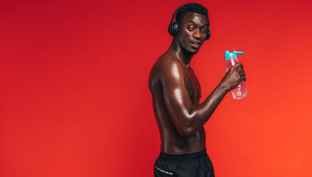 Afroamerikanischer Mann mit muskulösem Körper, der wegschaut, während er Wasser aus einer Flasche auf rotem Hintergrund trinkt. Ein fitter Mann hört Musik mit Kopfhörern während des Trainings. - JLPSF21685