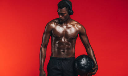 Fit junger Mann mit Medizinball vor rotem Hintergrund. Afrikanisches männliches Modell mit muskulösem Körper, das Kopfhörer trägt und einen Fitnessball in der Hand hält. - JLPSF21686