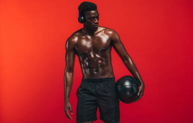 Mann ohne Hemd steht mit Medizinball vor rotem Hintergrund. Afroamerikanischer Mann mit muskulösem Körper hält einen Fitnessball und schaut weg. - JLPSF21687