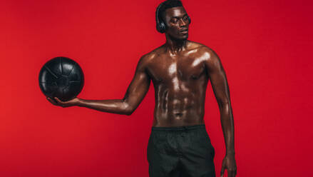 Kräftiger junger Mann mit muskulösem Körperbau, der einen Medizinball vor rotem Hintergrund hält und wegschaut. Afroamerikanischer Mann, der mit einem Fitnessball trainiert. - JLPSF21688