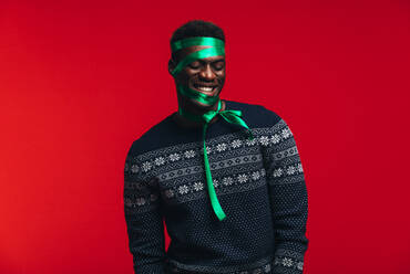 Glücklicher afrikanischer Mann mit Satinband auf seinem Gesicht vor rotem Hintergrund. Mann als Geschenk verpackt. - JLPSF21698