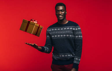Porträt eines jungen schwarzen Mannes, der ein Geschenk in die Höhe wirft. Afroamerikanischer Mann in Freizeitkleidung mit einer Geschenkbox auf rotem Hintergrund. - JLPSF21705