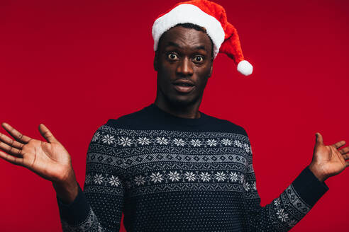 Afroamerikanischer Mann mit Weihnachtsmannmütze auf rotem Hintergrund, der überrascht in die Kamera blickt. - JLPSF21713