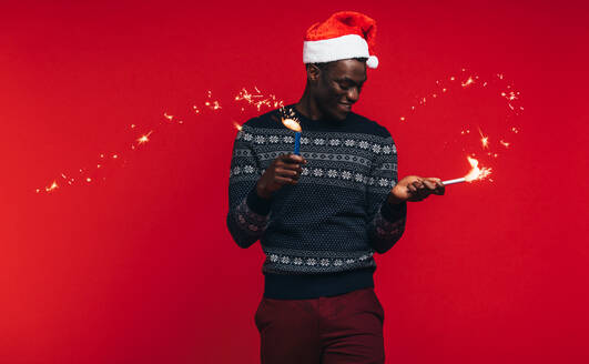 Junger Mann mit Weihnachtsmannmütze und Wunderkerzen auf rotem Hintergrund. Junger Mann genießt Weihnachten mit Feuerwerk. - JLPSF21719