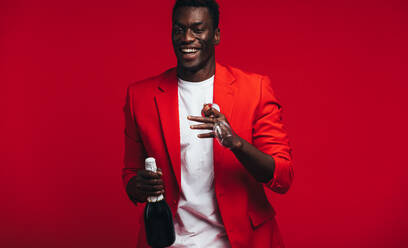 Fröhlicher junger afrikanischer Mann mit Champagnerflasche und Glas. Gutaussehender Mann in roter Jacke mit Champagner und Glas vor rotem Hintergrund. - JLPSF21722