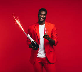 Gutaussehender junger afrikanischer Mann mit Sektflasche, Glas und Feuerwerk in der Hand. Stilvoller Mann im roten Anzug feiert Silvester vor rotem Hintergrund. - JLPSF21723
