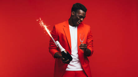 Afrikanischer Mann feiert das neue Jahr mit Champagner und Feuerwerk. Stilvoller Mann hat Spaß vor rotem Hintergrund. - JLPSF21724