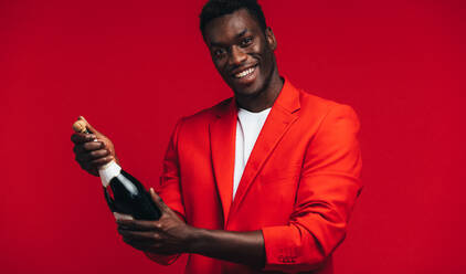 Hübscher junger afrikanischer Mann, der eine Champagnerflasche vor rotem Hintergrund öffnet. Schwarzer Mann, der mit Champagner feiert. - JLPSF21725