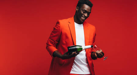Hübscher junger afrikanischer Mann gießt Champagner in ein Glas. Lächelnder junger Mann mit Champagner vor rotem Hintergrund. - JLPSF21726