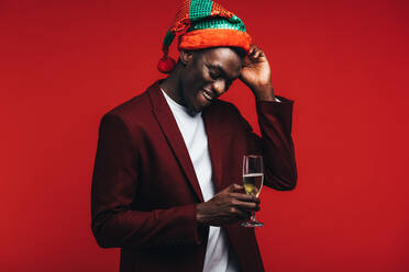 Young man wearing a party hat standing on red background with a glass of champagne. African american man enjoying at party with drinks. - JLPSF21735