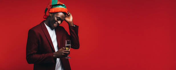 Young man wearing a party hat standing on red background with a glass of champagne. African american man enjoying at party with drinks. - JLPSF21736