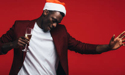 African man wearing santa hat with champagne dancing against red background. Happy young black guy enjoying at christmas party. - JLPSF21737