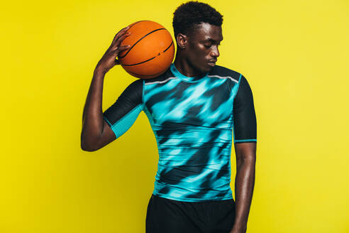 Afrikanischer Mann mit Basketball auf der Schulter, männlicher Basketballspieler vor gelbem Hintergrund. - JLPSF21752