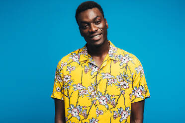 Portrait of african man wearing a Hawaiian style floral shirt and looking at camera. Young guy in summer style outfit against blue background. Tropical holiday concept. - JLPSF21760