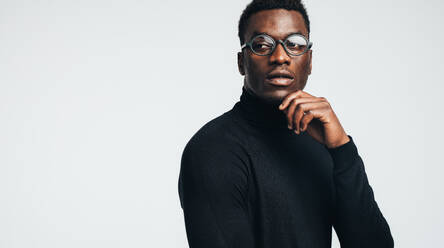 Gut aussehender junger afrikanischer Mann in schwarzem Polo-T-Shirt und Brille, der vor einem grauen Hintergrund wegschaut. - JLPSF21766