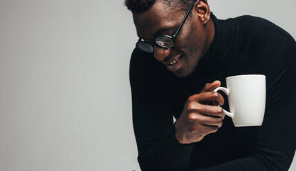 African man with cup of coffee looking down and smiling. Man relaxing with a coffee against gray background. - JLPSF21774