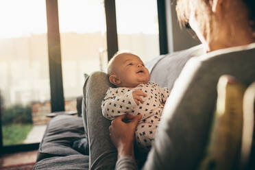 Blick über die Schulter einer Mutter, die mit ihrem Kleinkind auf einer Couch zu Hause sitzt. Rückansicht einer Mutter, die mit ihrem Baby spielt, mit Sonnenfackel im Hintergrund. - JLPSF21785