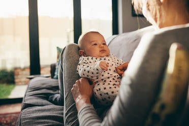 Rückansicht einer Mutter, die zu Hause auf der Couch sitzt und ihr Baby hält. Frau sitzt zu Hause und spielt mit ihrem kleinen Kind. - JLPSF21786