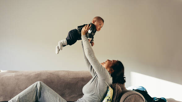 Junge Mutter spielt mit ihrem Baby, das zu Hause auf der Couch sitzt. Frau sitzt auf einem Sofa und hebt ihr Baby hoch. - JLPSF21788