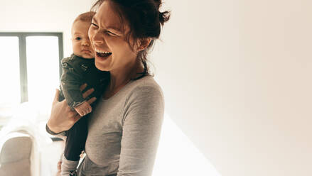 Nahaufnahme einer lächelnden Frau, die in ihrem Haus steht und ihr Kind trägt. Frau, die ihr Baby hält und lachend im Raum steht. - JLPSF21793