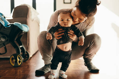 Nahaufnahme einer Frau, die mit ihrem Baby spielt, das auf dem Boden hockt und gehalten wird. Mutter lässt ihr Baby zu Hause auf dem Boden stehen. - JLPSF21798