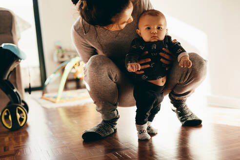 Nahaufnahme einer Frau, die mit ihrem Baby spielt, das auf dem Boden hockt und gehalten wird. Mutter lässt ihr Baby zu Hause auf dem Boden stehen. - JLPSF21801