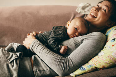 Smiling mother lying on couch with her baby sleeping on her chest. Happy mother relaxing at home with her child sleeping on her. - JLPSF21802