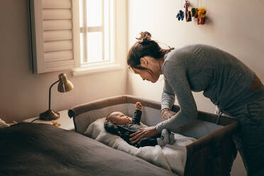 Mother putting her baby to sleep on a bedside baby crib. Woman bending forward over a crib to check her sleeping baby. - JLPSF21808