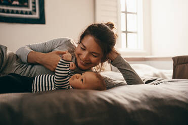 Frau auf dem Bett liegend, die ihr Baby verwöhnt. Lächelnde Mutter, die mit ihrem Kleinkind spielt, das zu Hause auf dem Bett liegt. - JLPSF21816