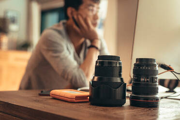 Two professional camera lenses and portable hard disk on table with photographer working on computer at the back. Photographer editing photos on desktop with camera lenses placed on table. - JLPSF21817