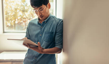 Asiatischer Mann lehnt sich an eine Wand und benutzt ein digitales Tablet. Ein Mann in Freizeitkleidung steht zu Hause und benutzt einen Tablet-Computer, um seinen Status in den sozialen Medien zu aktualisieren. - JLPSF21832