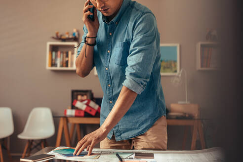 Designer talking on mobile phone while choosing color for interior in office. Asian man looking at color palette and taking with client over phone. - JLPSF21836