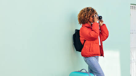 Weibliche afroamerikanische Touristin, die mit ihrem Gepäck im Freien steht und Fotos mit einer Digitalkamera schießt. Frau im Urlaub, die im Freien vor einer Wand stehend ein Foto macht. - JLPSF21845