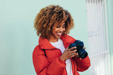 Fröhliche afroamerikanische Frau im Urlaub, die im Freien stehend Fotos macht. Porträt einer Touristin, die auf ihre DSLR-Kamera schaut, nachdem sie ein Foto gemacht hat. - JLPSF21846