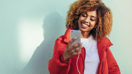 Fröhliche afroamerikanische Frau, die auf ihr Mobiltelefon schaut und einen Videochat führt. Lächelnde Frau, die ein Selfie mit ihrem Mobiltelefon macht, während sie Musik hört. - JLPSF21848