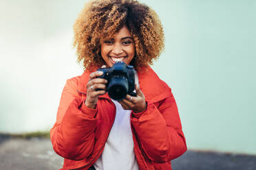 Porträt einer fröhlichen afroamerikanischen Frau, die eine DSLR-Kamera hält. Weibliche Reisende, die mit einer Digitalkamera Fotos im Freien schießt. - JLPSF21852