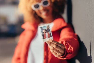 Unscharfe Aufnahme einer weiblichen Reisenden im Urlaub, die ein Polaroid-Foto zeigt. Nahaufnahme eines Polaroid-Fotos in der Hand einer lächelnden weiblichen Reisenden, die im Freien steht. - JLPSF21854