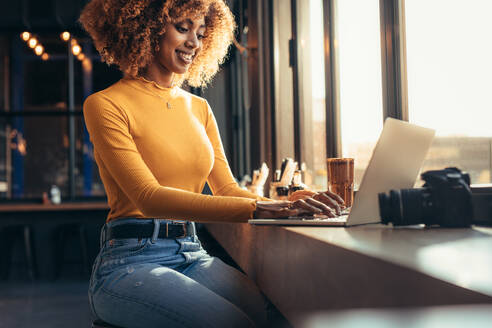 Freiberuflicher Fotograf sitzt in einem Restaurant in der Nähe eines Fensters und arbeitet an einem Laptop. Lächelnder afroamerikanischer Blogger arbeitet an einem Laptop mit einer Digitalkamera vor sich. - JLPSF21857